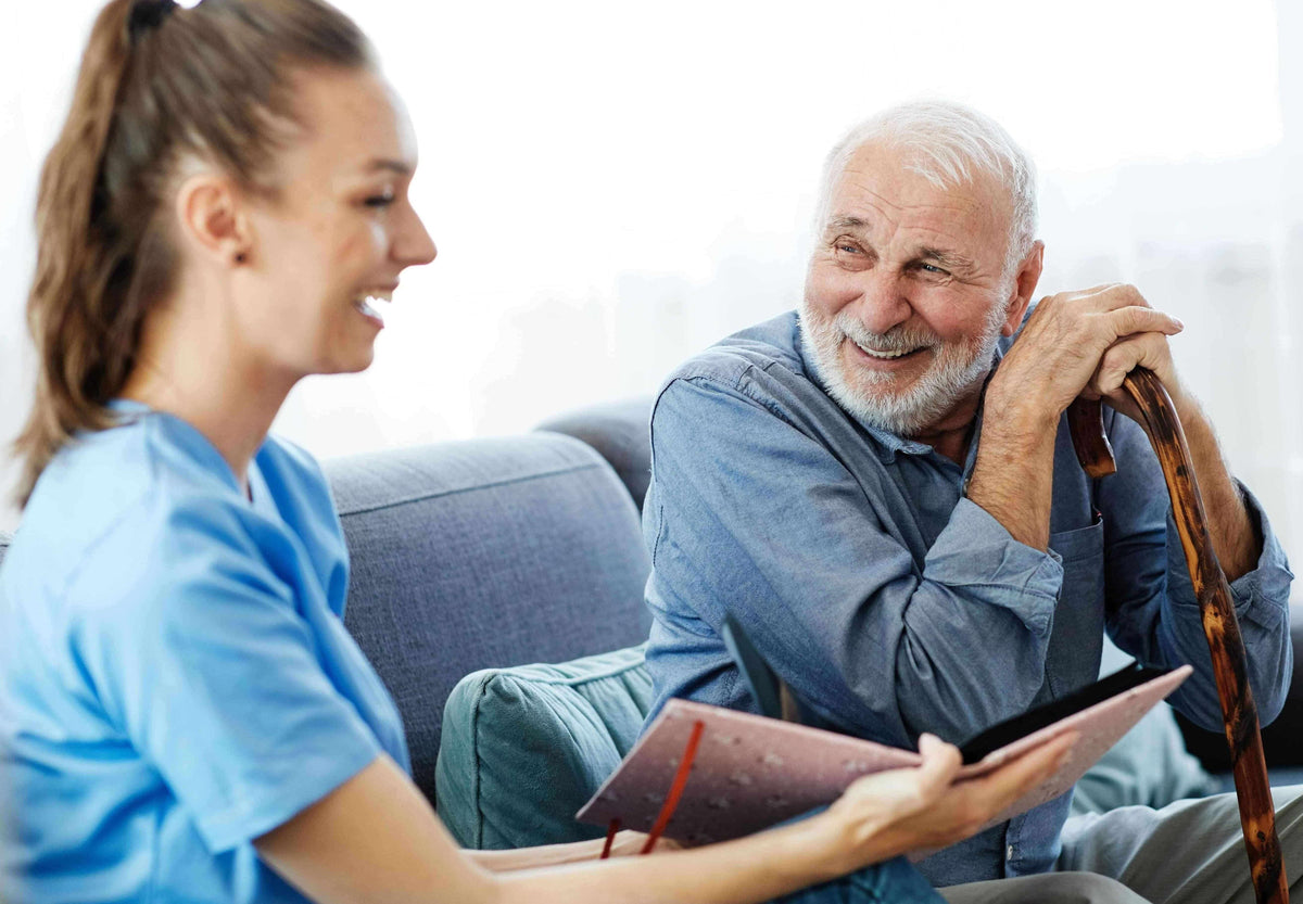 Older veteran smiling while talking with a home health nurse on the couch, clipboard in hand.