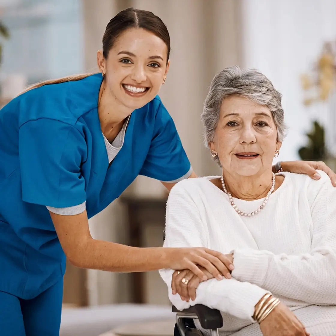 A smiling female healthcare worker in blue scrubs leans over an elderly woman seated in a wheelchair, offering support in a bright indoor care setting.