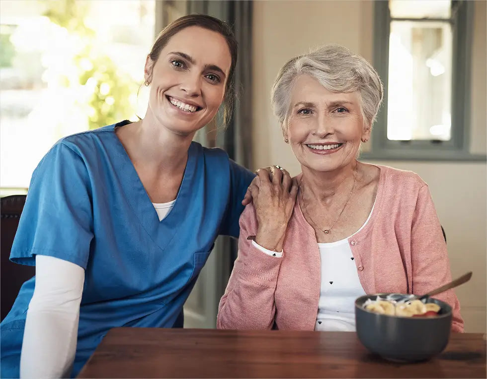 A smiling healthcare worker in blue scrubs sits beside an elderly woman at a table, with the older woman wearing a pink cardigan and a bowl of food in front of them.
