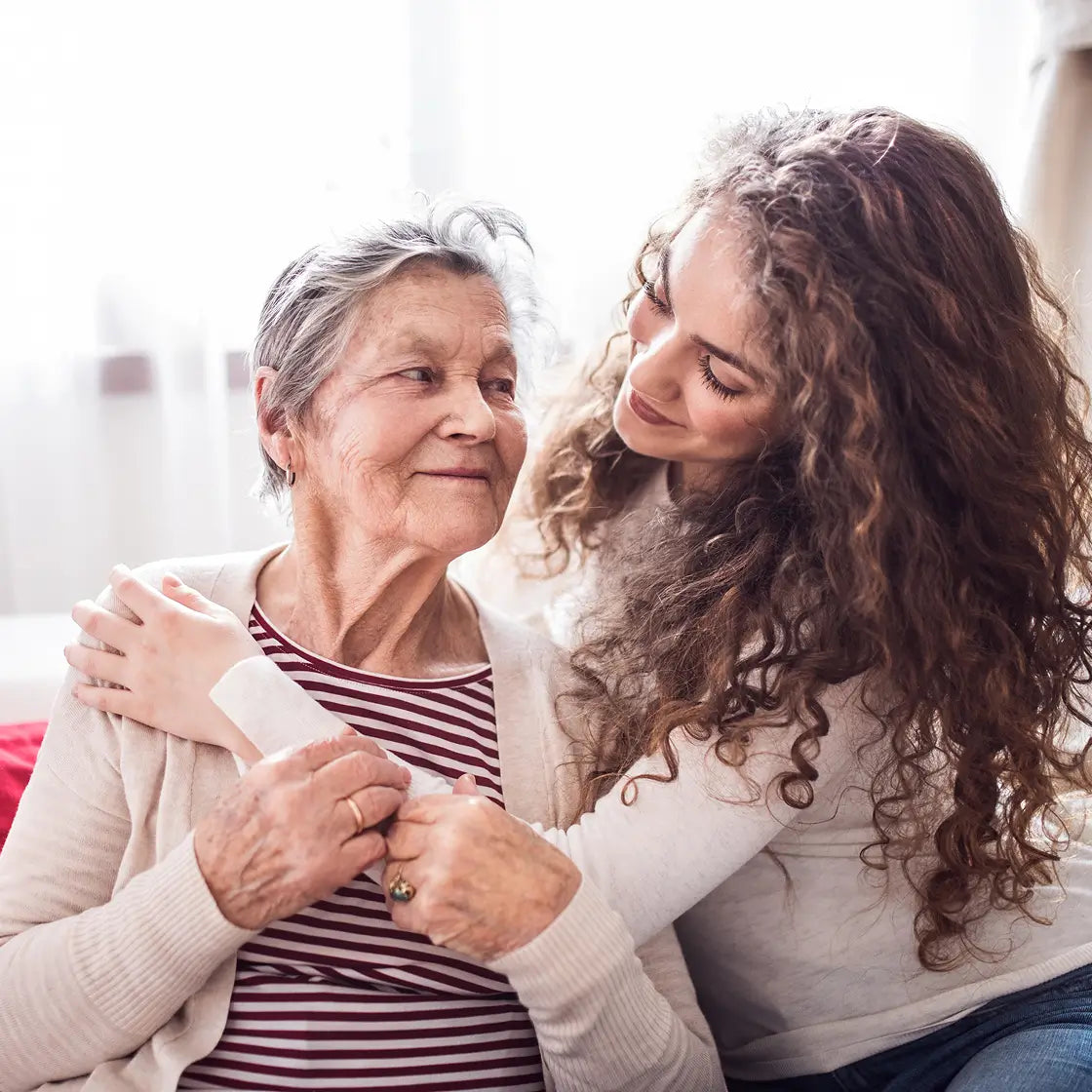 An elderly woman with gray hair sits smiling while a younger woman with long curly brown hair leans in close and comforts her. The younger woman wears a light beige top, and the older woman wears a cream cardigan over a red-and-white striped shirt.