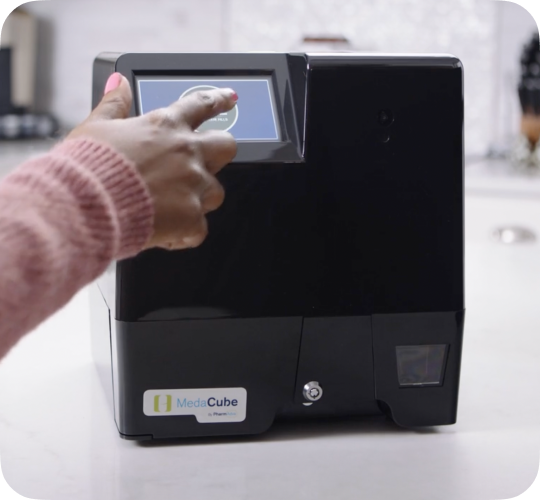 A MedaCube sitting on a kitchen countertop with a figer about the press the screen to dispense medications.