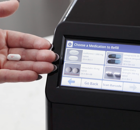 Hand holding a pill next to the MedaCube screen which has the refill menu open showing 4 of the different medications loaded into the MedaCube.