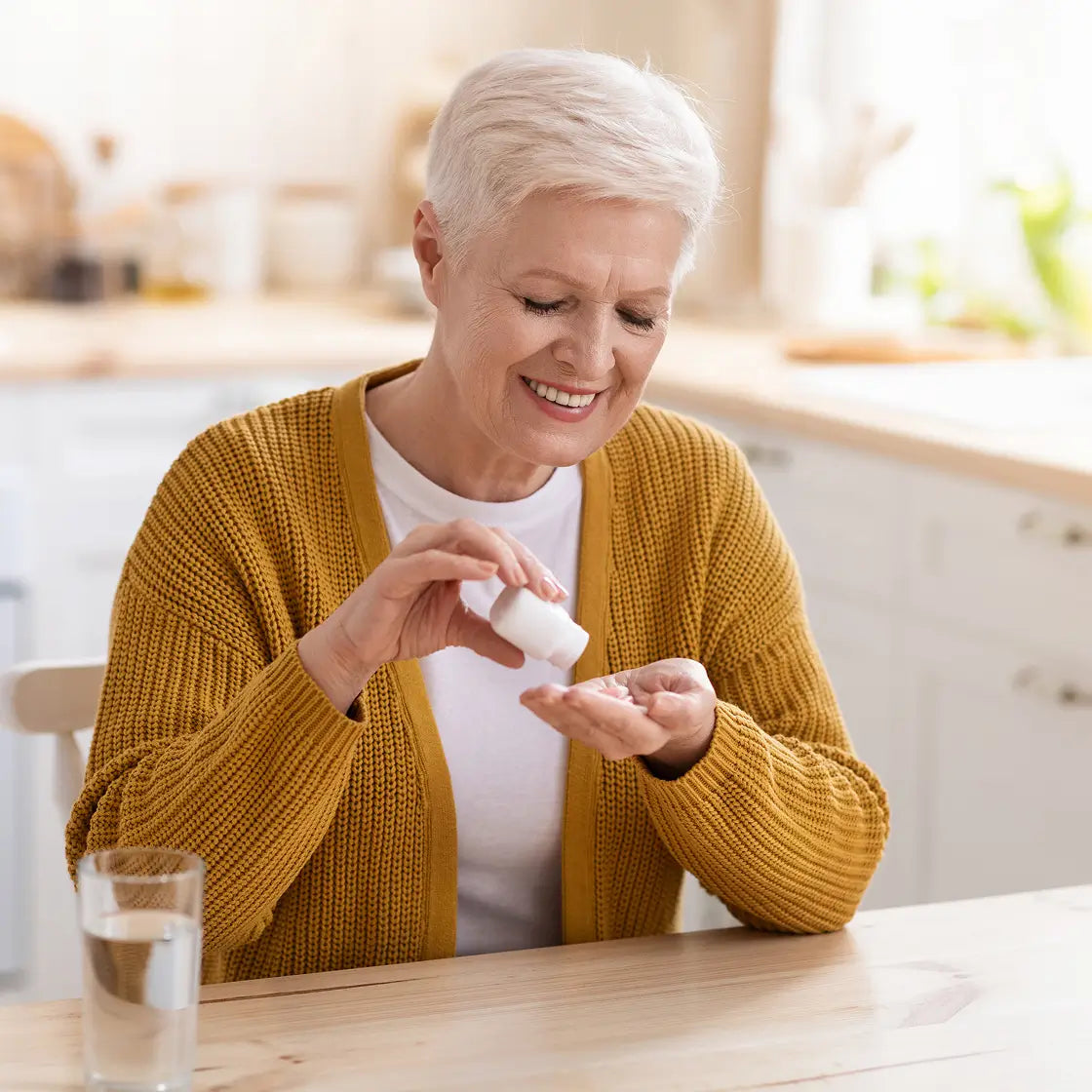 An older woman with short white hair smiles while holding a small white pill bottle above her palm, with a glass of water on the table in a bright kitchen setting.