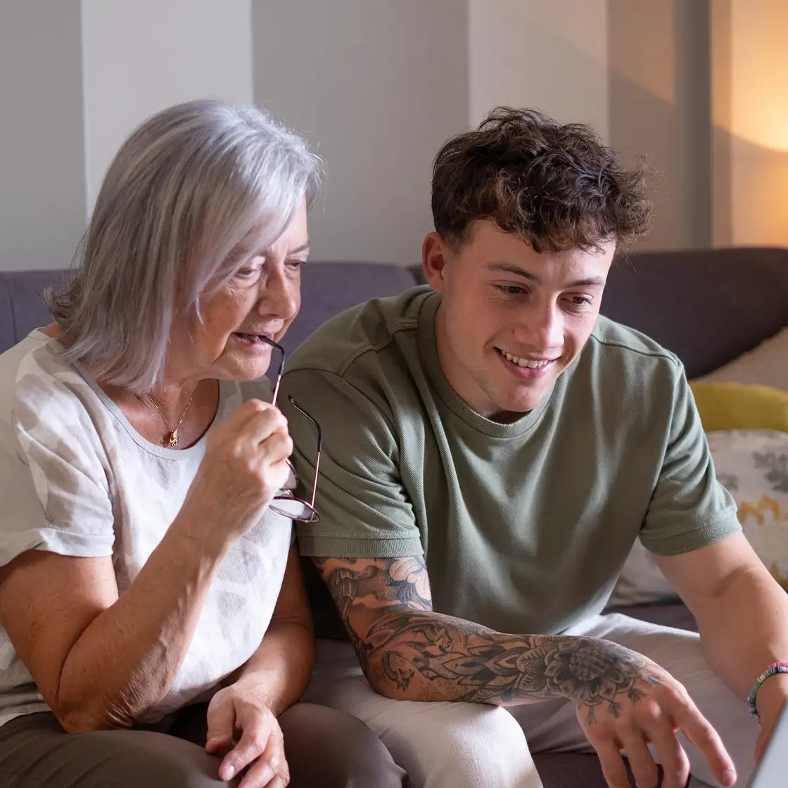 An older woman with gray hair and a young man with curly brown hair sitting together on a couch in a living room. The woman is wearing a white t-shirt and holding a pair of eyeglasses, while the man wears an olive green t-shirt and light-colored pants.