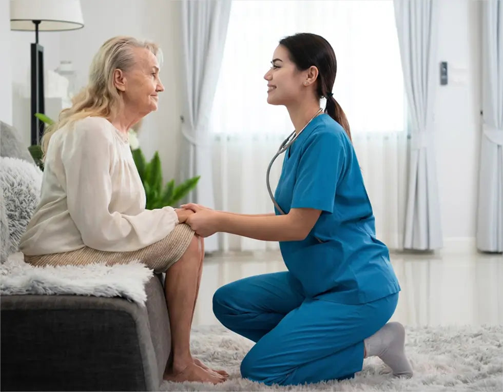 A female healthcare worker in blue scrubs kneels on a white rug, holding hands with an elderly woman sitting on a gray sofa in a bright, modern room.