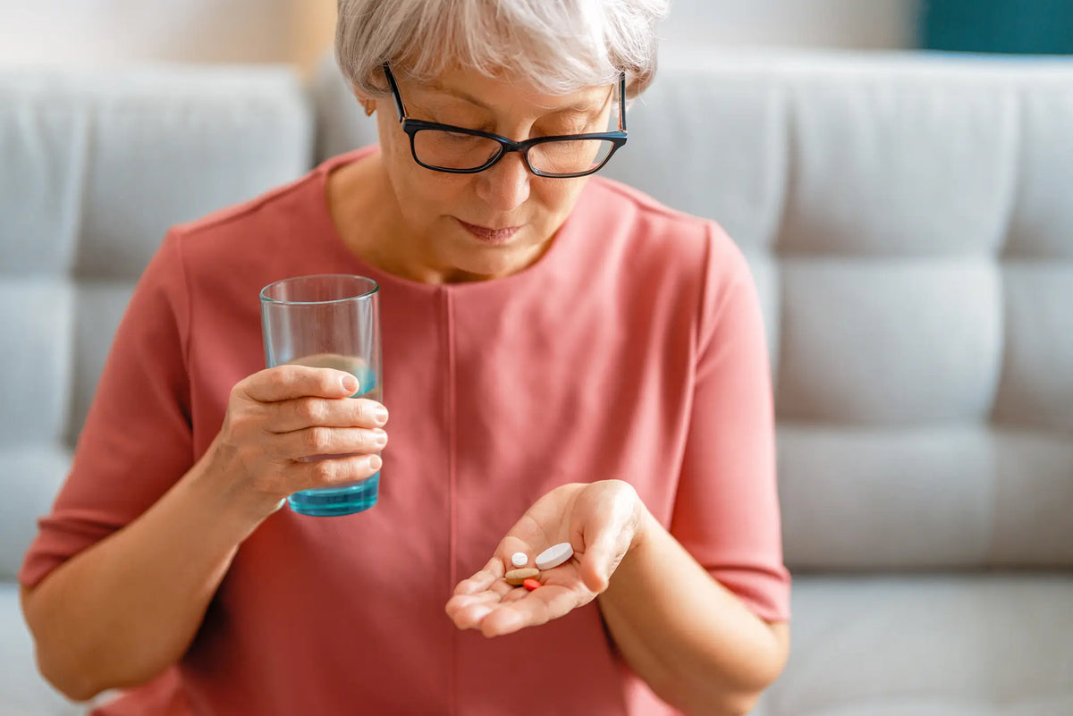 An older woman with short gray hair and black-rimmed glasses looks down at white and orange pills in her palm while holding a clear glass of water. She is wearing a dusty rose pink top and sitting in front of a light gray tufted sofa.